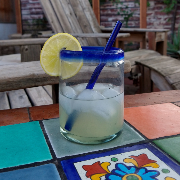 Daiquiri cocktail in a clear glass with a blue rim and a blue straw, sitting on a colorful tile table in front of a large yard with wooden lounge chairs