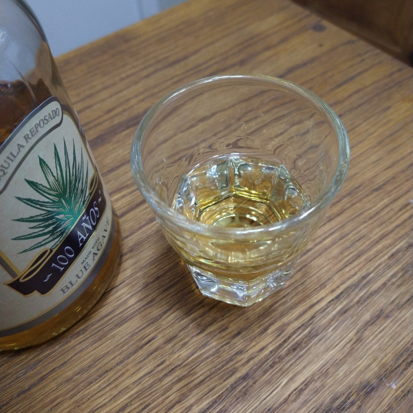 Small glass of golden-yellow tequila, next to a bottle of 100 Años reposado, shot at an angle so only the label is showing.
