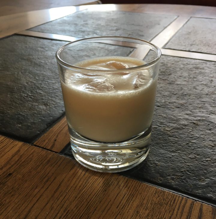 Light beige colored beverage with ice in a clear glass, on top of a dark grey stone table