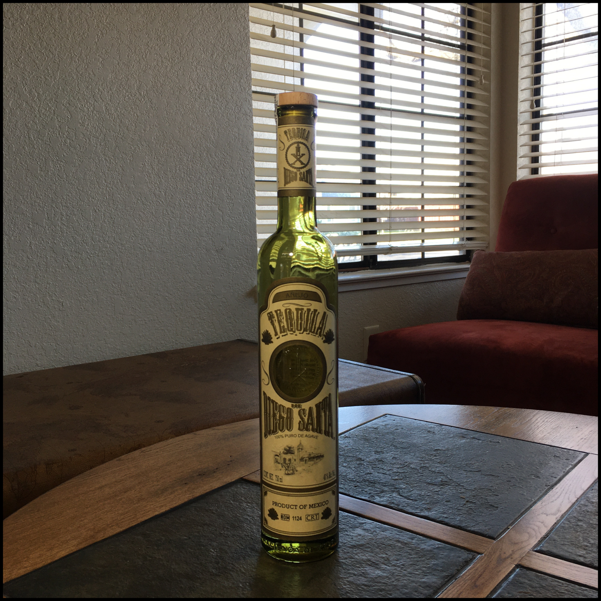 Bottle of Don Diego Santa Tequila Añejo on top of a grey stone and wooden table, in front of a living room window and background furniture.