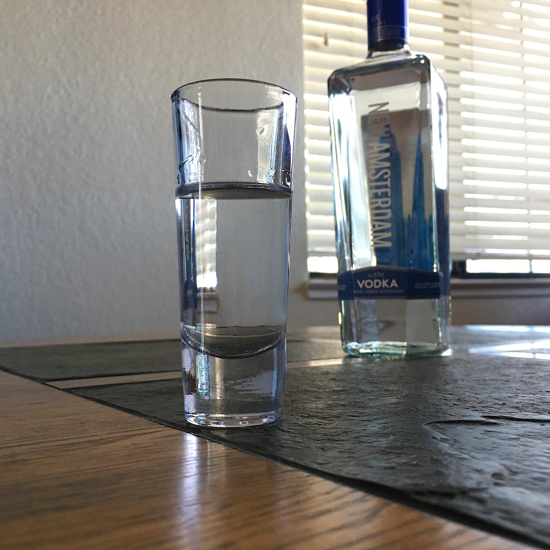 Shot glass full of New Amsterdam Vodka on top of a stone and wood table, with the vodka bottle in the background