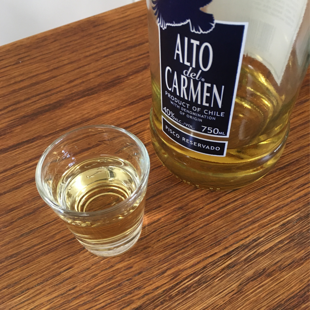 Shot glass full of Alto del Carmen Pisco Reservado, next to the bottle which has the label partially showing, on top of a wooden table