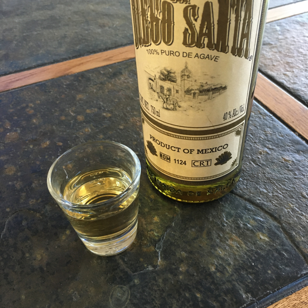 Clear shot glass full of the golden colored Don Diego Santa Añejo Tequila, next to the tequila bottle with the label partially showing, on top of a dark grey stone table