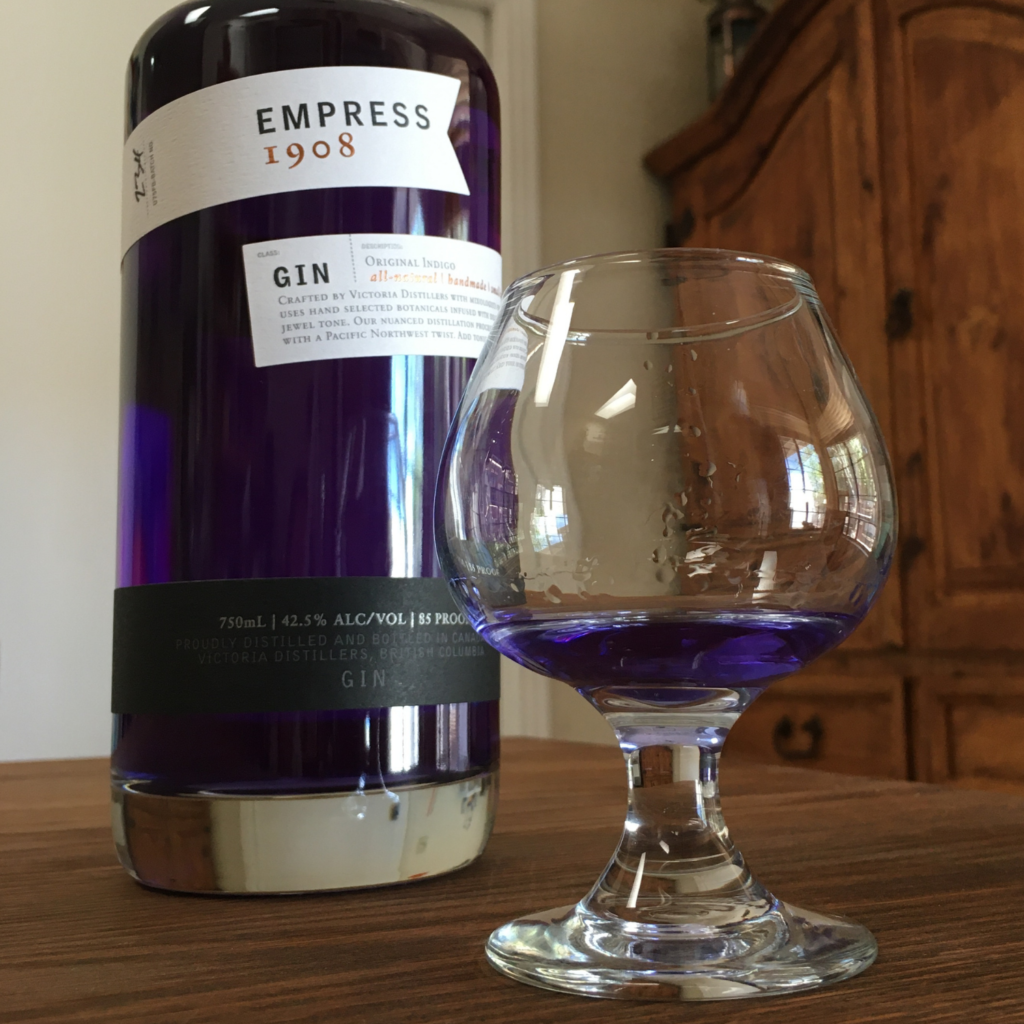 Round glass with indigo liquid, next to a bottle of Empress 1908 Gin, on a dark brown table and in front of a mixed white and wooden background