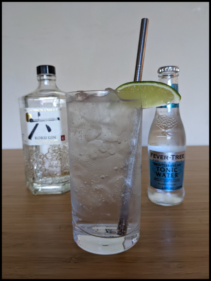 glass of ice and clear liquid with a green lime and metal straw with a clear Roku Gin bottle with blue label and clear bottle with white label on a light brown table with white background