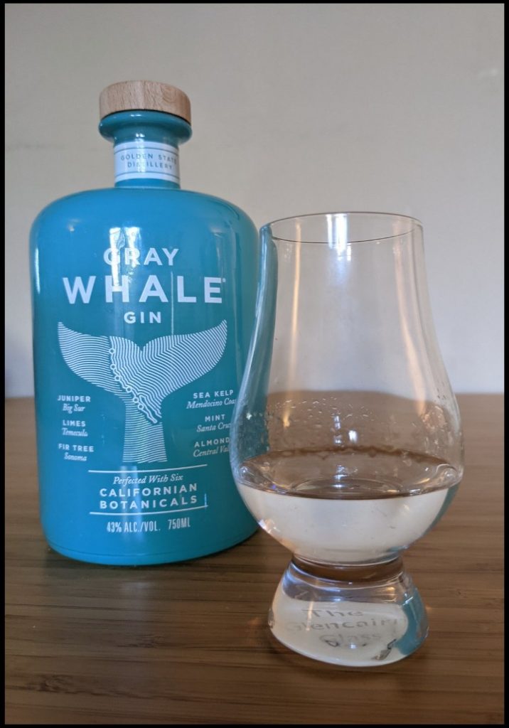clear liquid in a glencairn glass on a light brown table with a blue bottle of Gray Whale Gin behind it and a white background