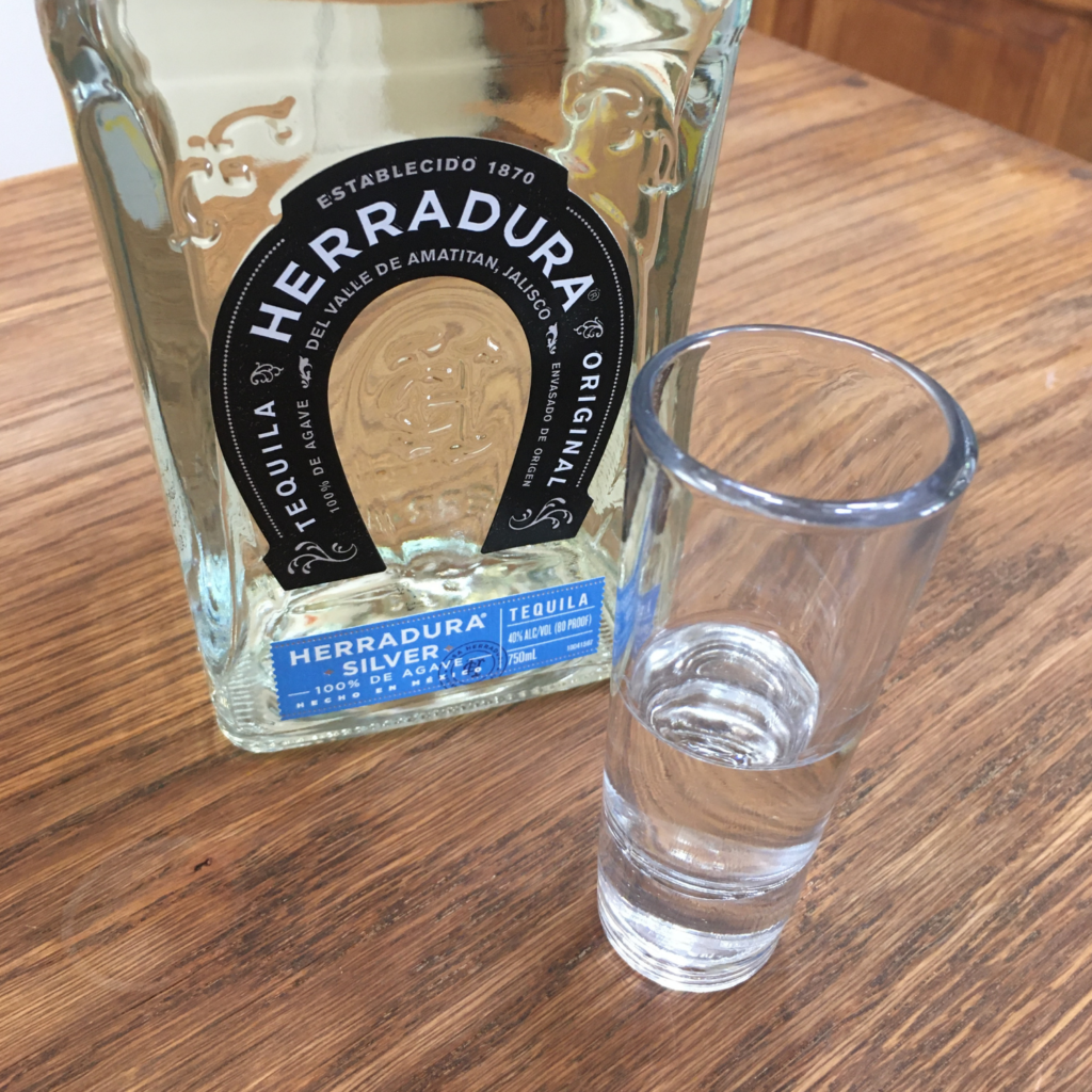 Tall shot glass with clear liquid next to a bottle of Herradura Silver Tequila with part of the bottle showing, on a wooden table