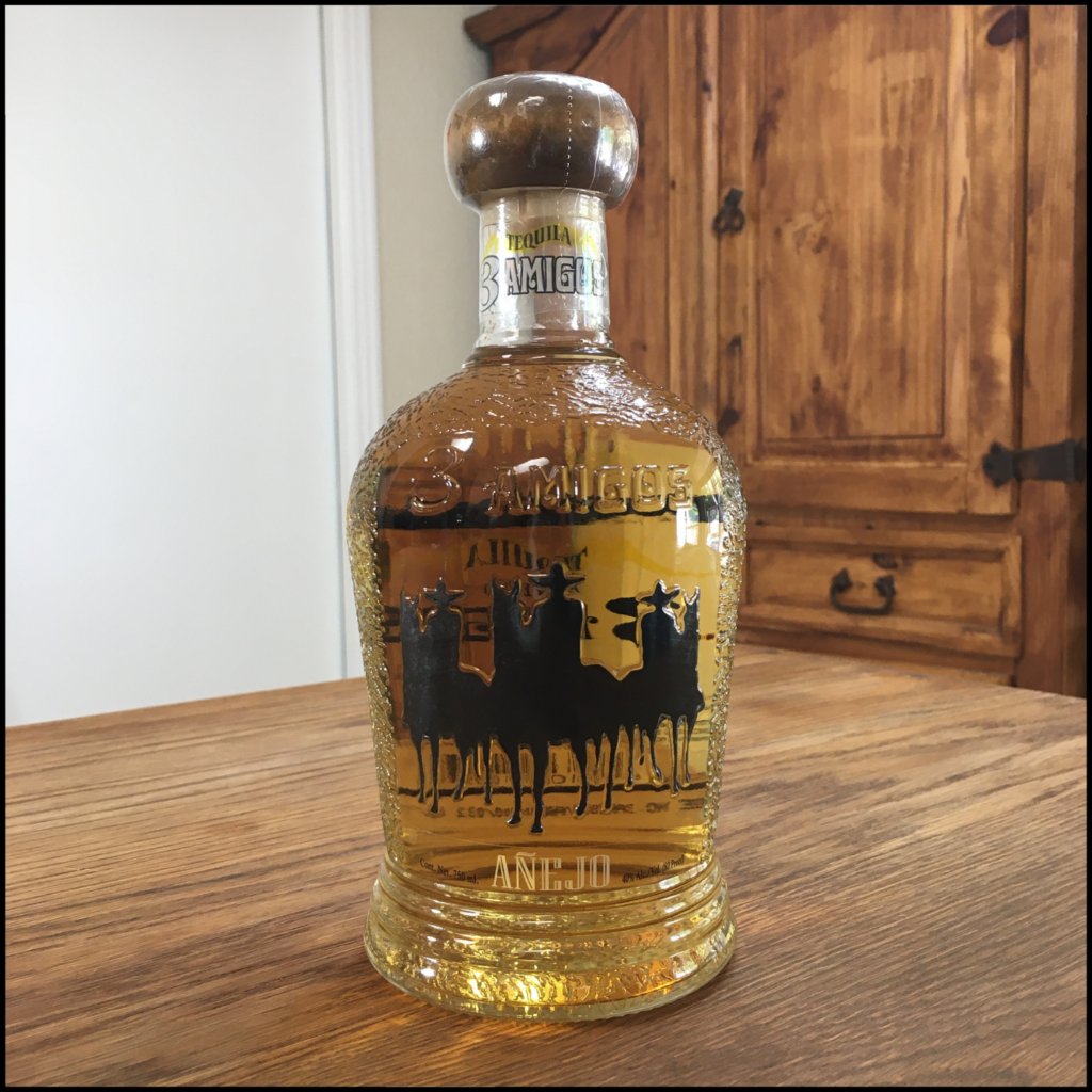 Bottle of 3 Amigos Tequila Añejo sittingon a wooden table, in front of a mixed white and wooden background