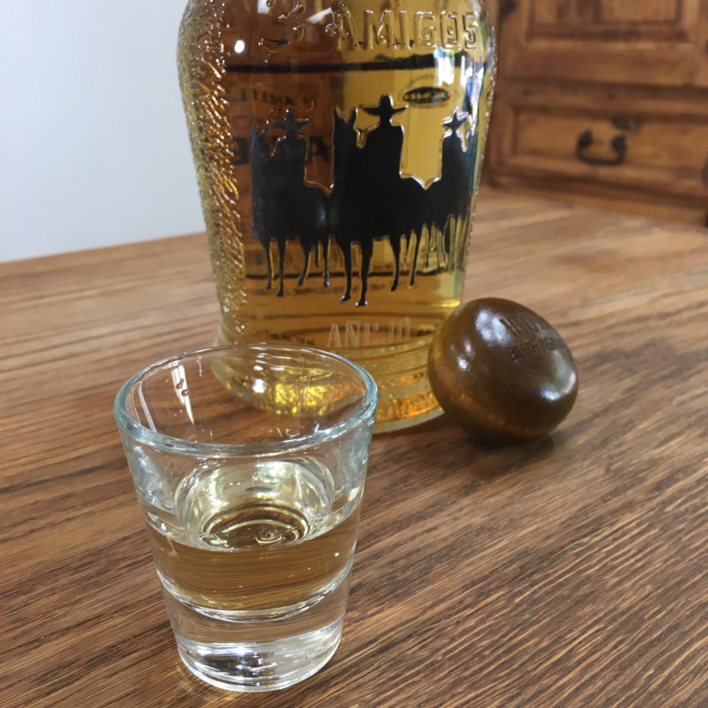 Shot glass full of golden yellow liquid, next to an open bottle of 3 Amigos Tequila Añejo, on a wooden table top