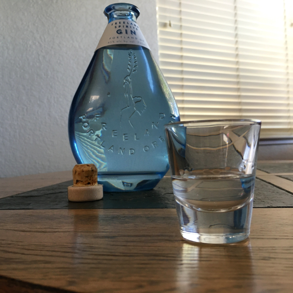 Shot glass filled with clear liquid, in front of an open bottle of Freeland Spirits Gin, sitting on a mixed wooden and stone table