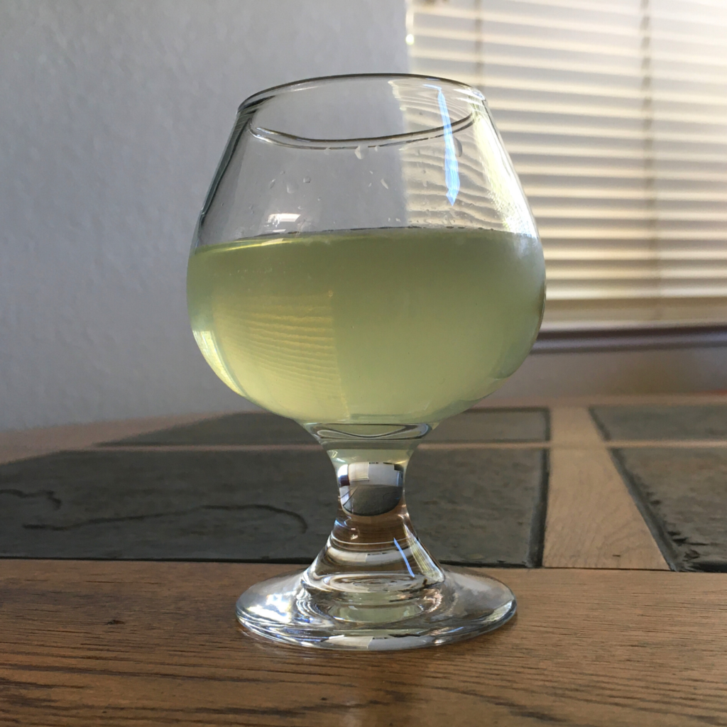 Soft yellow-green cocktail in a rounded cocktail glass, sitting on a mixed wood and stone table