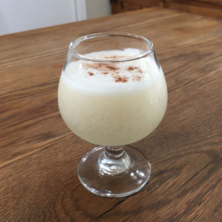 Off-white cocktail with a cinnamon garnish inside of a rounded cocktail glass, sitting on a wooden table top