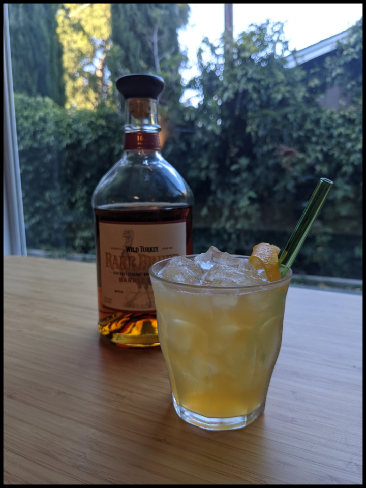 yellow liquid with ice in a old fashion glass with a lemon garnish and green glass straw with a whiskey bottle in the background