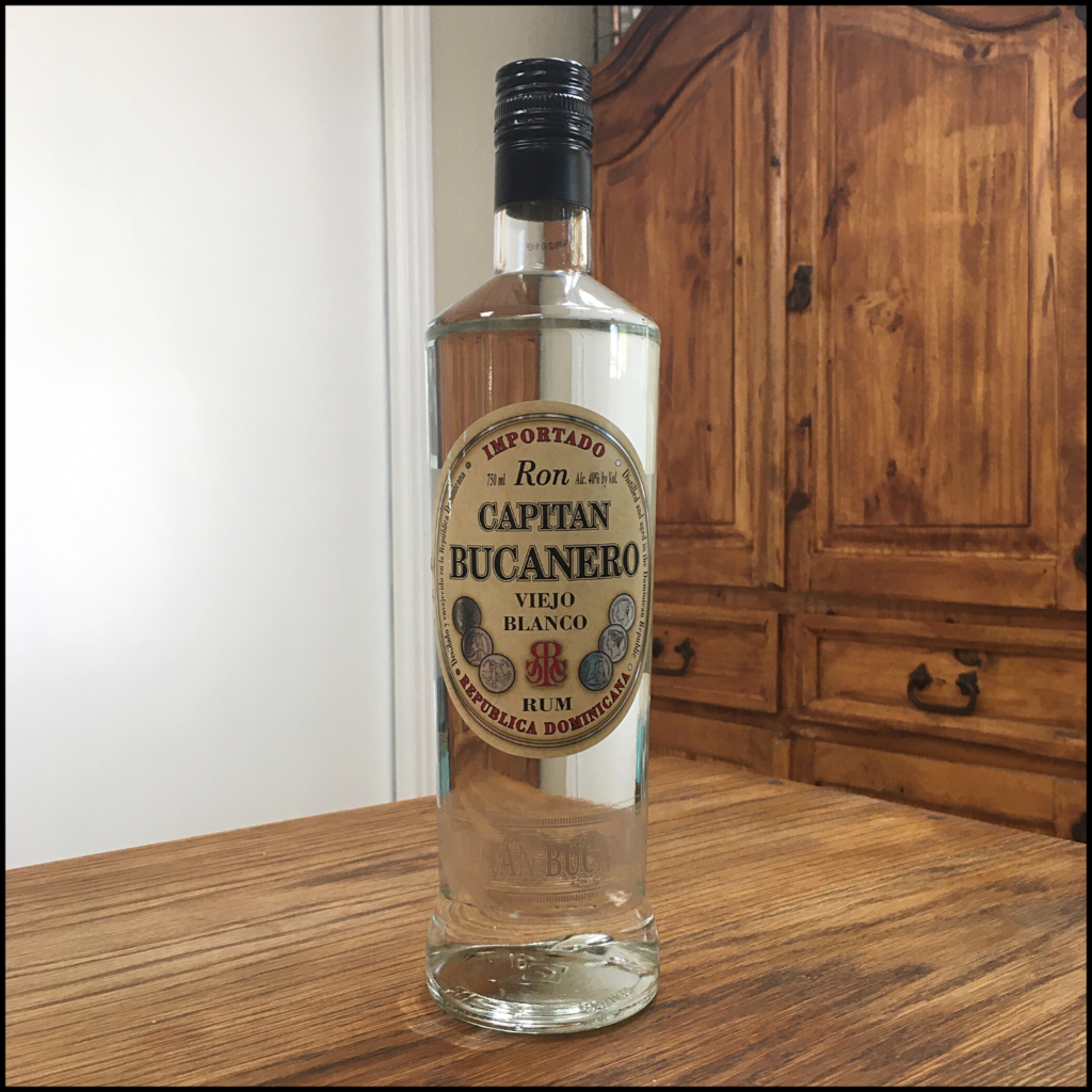 Bottle of Capitan Bucanero Viejo Blanco Rum, sitting on a wooden table in front of a mixed white and wooden background