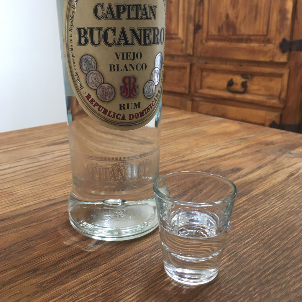 Shot glass with clear liquid next to a bottle of Capitan Bucanero Viejo Blanco rum, sitting on a wooden table in front of a mixed white and wooden background