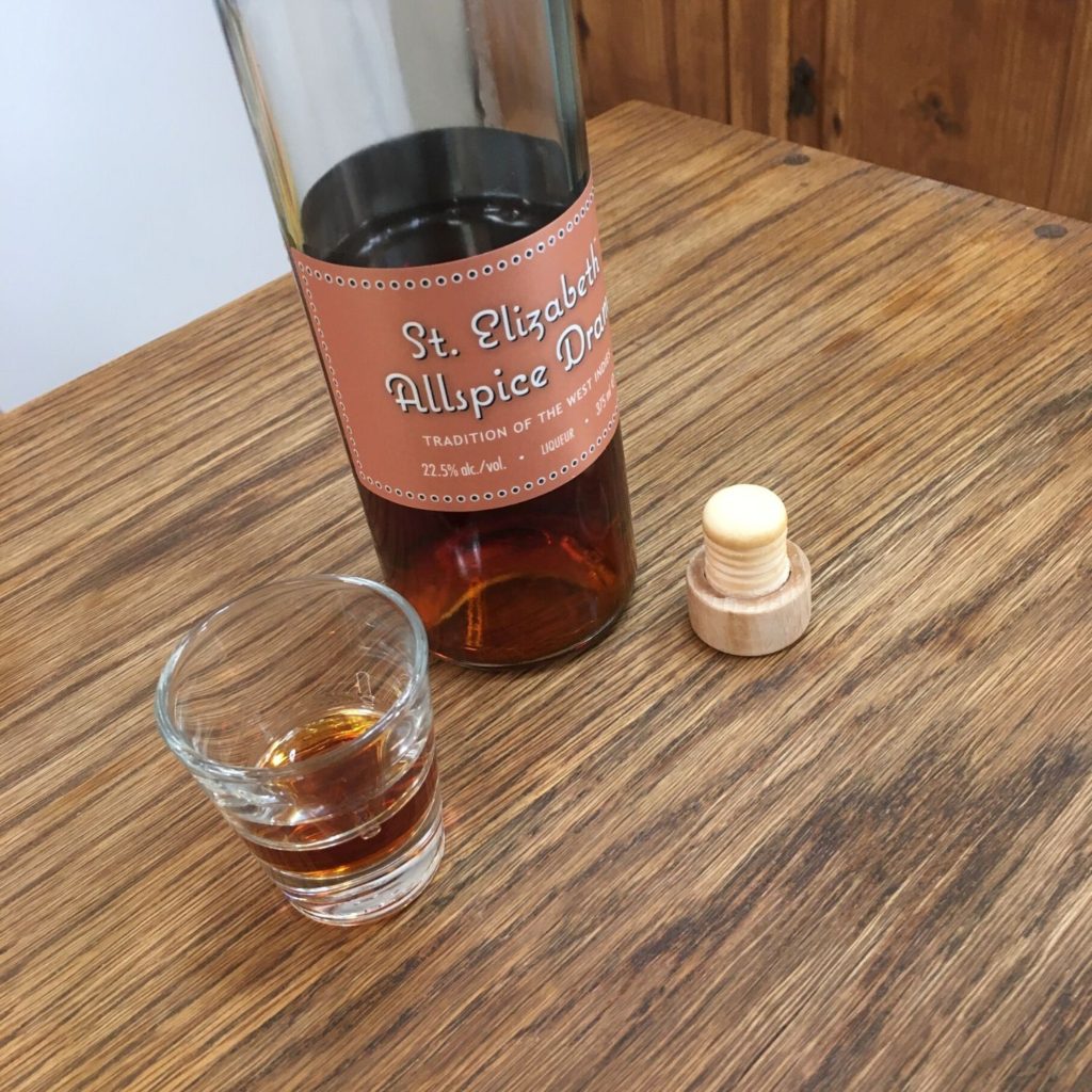 Shot glass half filled with dark amber liquid, next to a bottle of St. Elizabeth Allspice Dram and a cork top