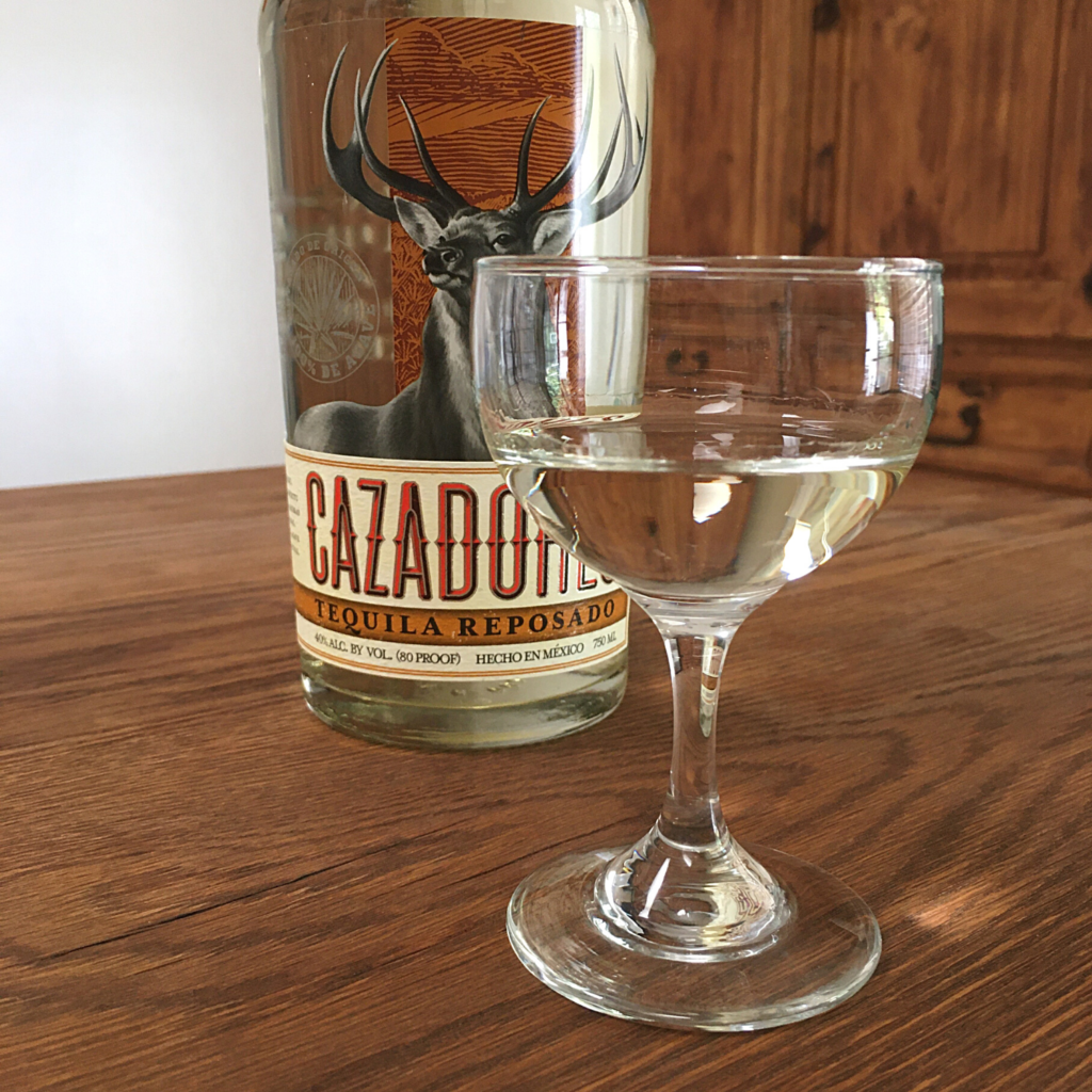 Small rounded glass with light golden liquid next to a bottle of Cazadores Tequila Reposado, sitting on a wooden table