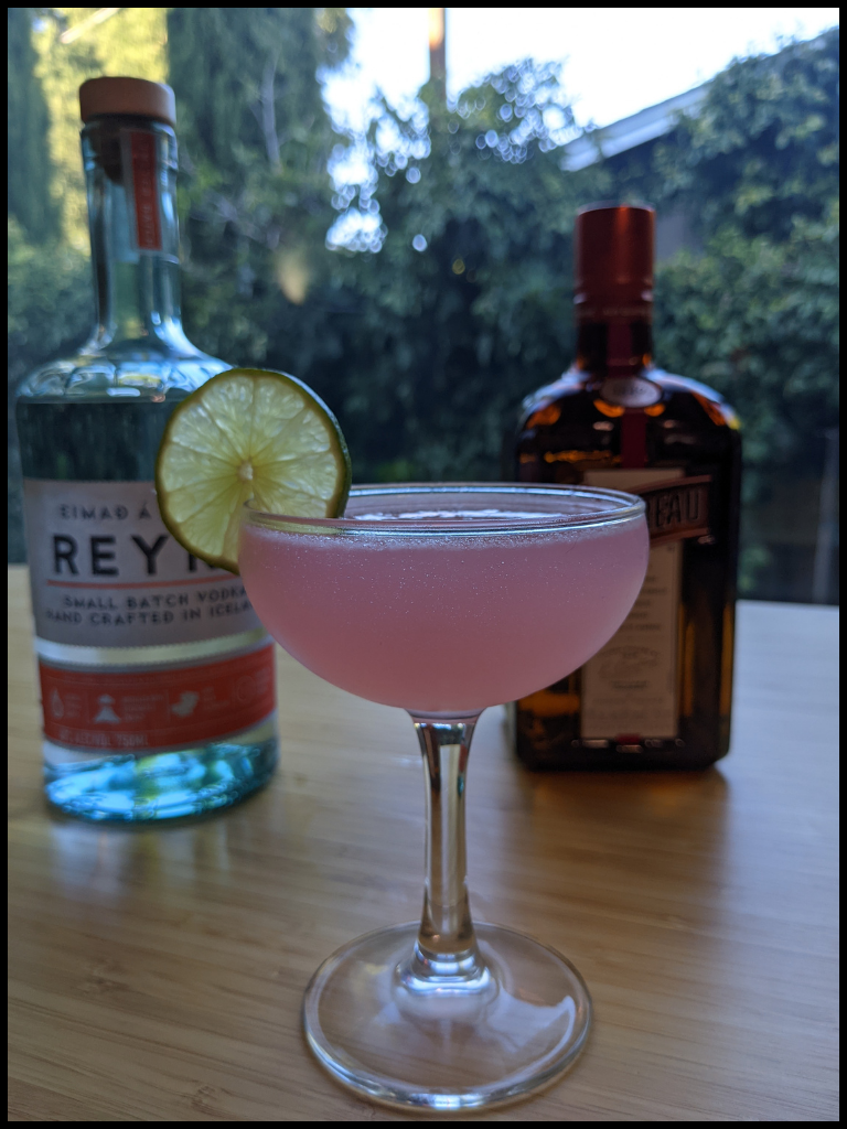 pink liquid in a coupe glass with a lime wheel with two alcohol bottles in the background