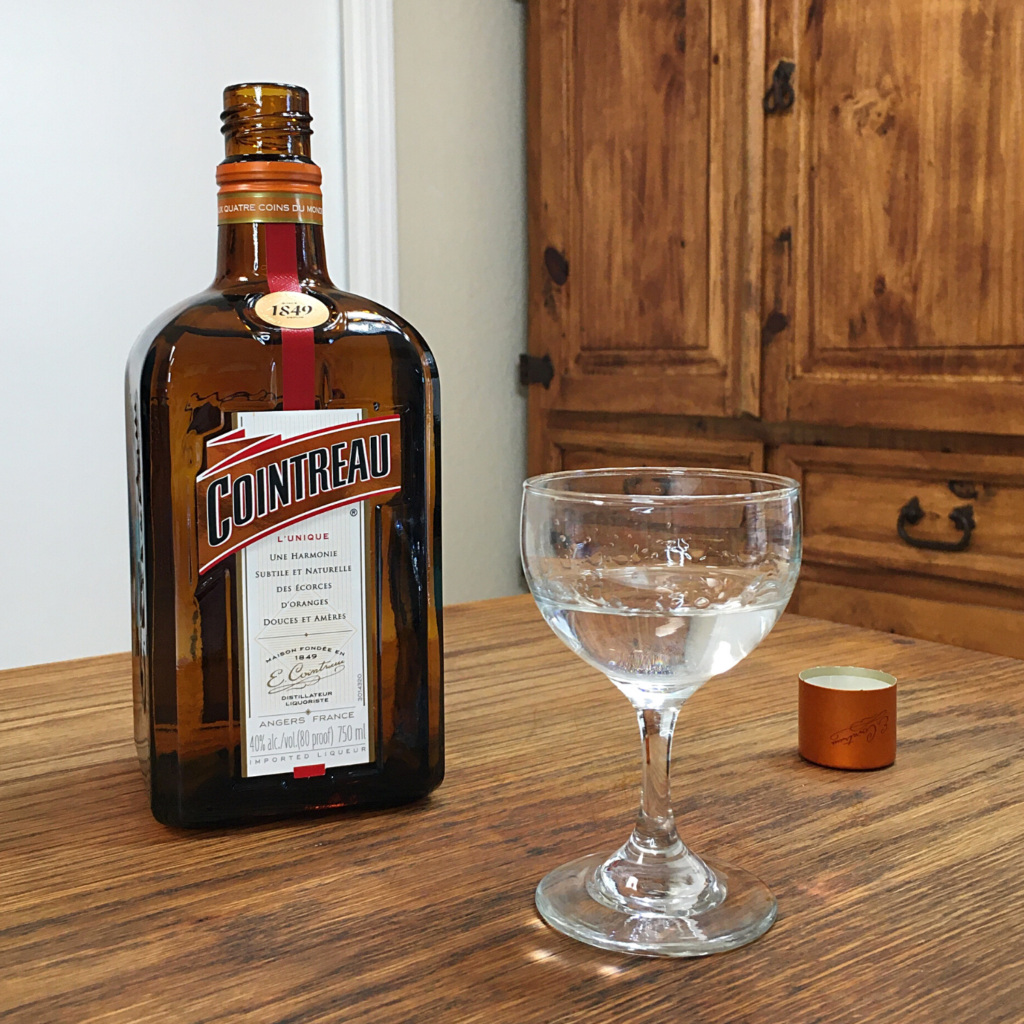 Open bottle of Cointreau orange liqueur, next to the cap and a rounded glass filled with clear liquid