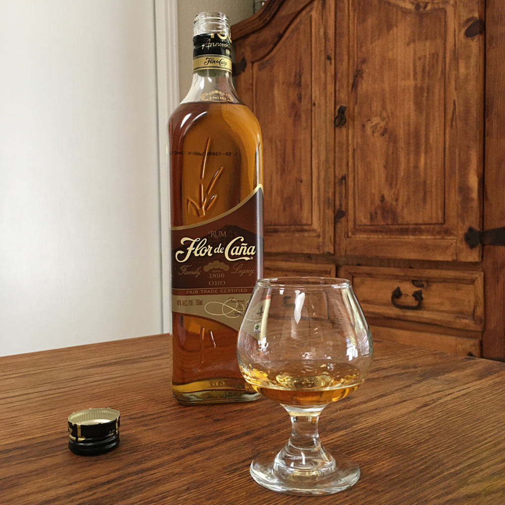 Open bottle of Flor de Caña 4 Añejo Oro rum, next to a round glass filled with golden liquid, sitting on a wooden table