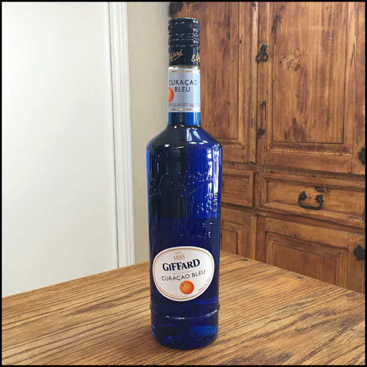 Bottle of Giffard Blue Curacao Liqueur sitting on a wooden table, in front of a mixed white and wooden background