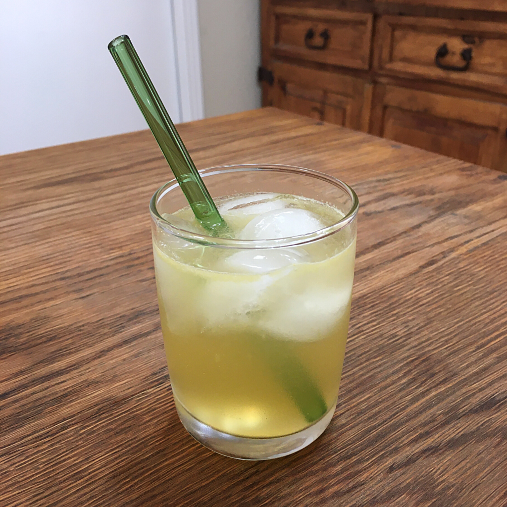 Yellow cocktail with ice and a deep green glass straw, sitting on a wooden table