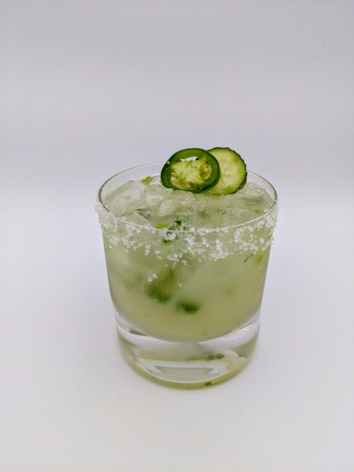 light green liquid with floating pieces of cumumber and jalapeno in a salted rocks glass with a slice of cucumber and jalapeno as a garnish