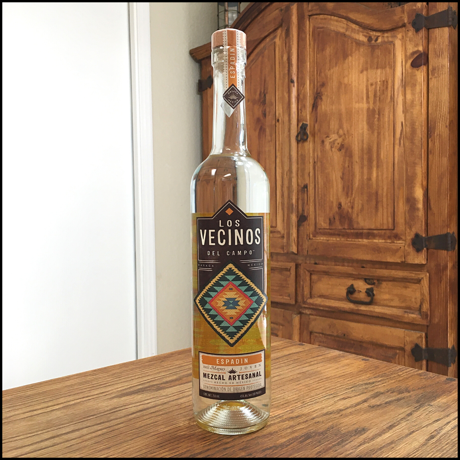 Bottle of clear-colored Los Vecinos del Campo Mezcal sitting on a wooden table, in front of a mixed white and wooden background