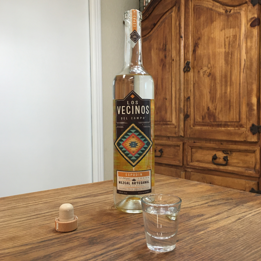 Shot glass filled with clear liquid next to an open bottle of Los Vecinos del Campo Mezcal, sitting on a wooden table