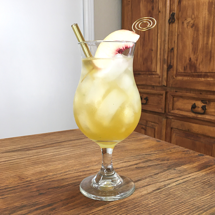 Yellow-colored cocktail with ice in a hurricane glass, with a yellow glass straw and white peach wedge garnish