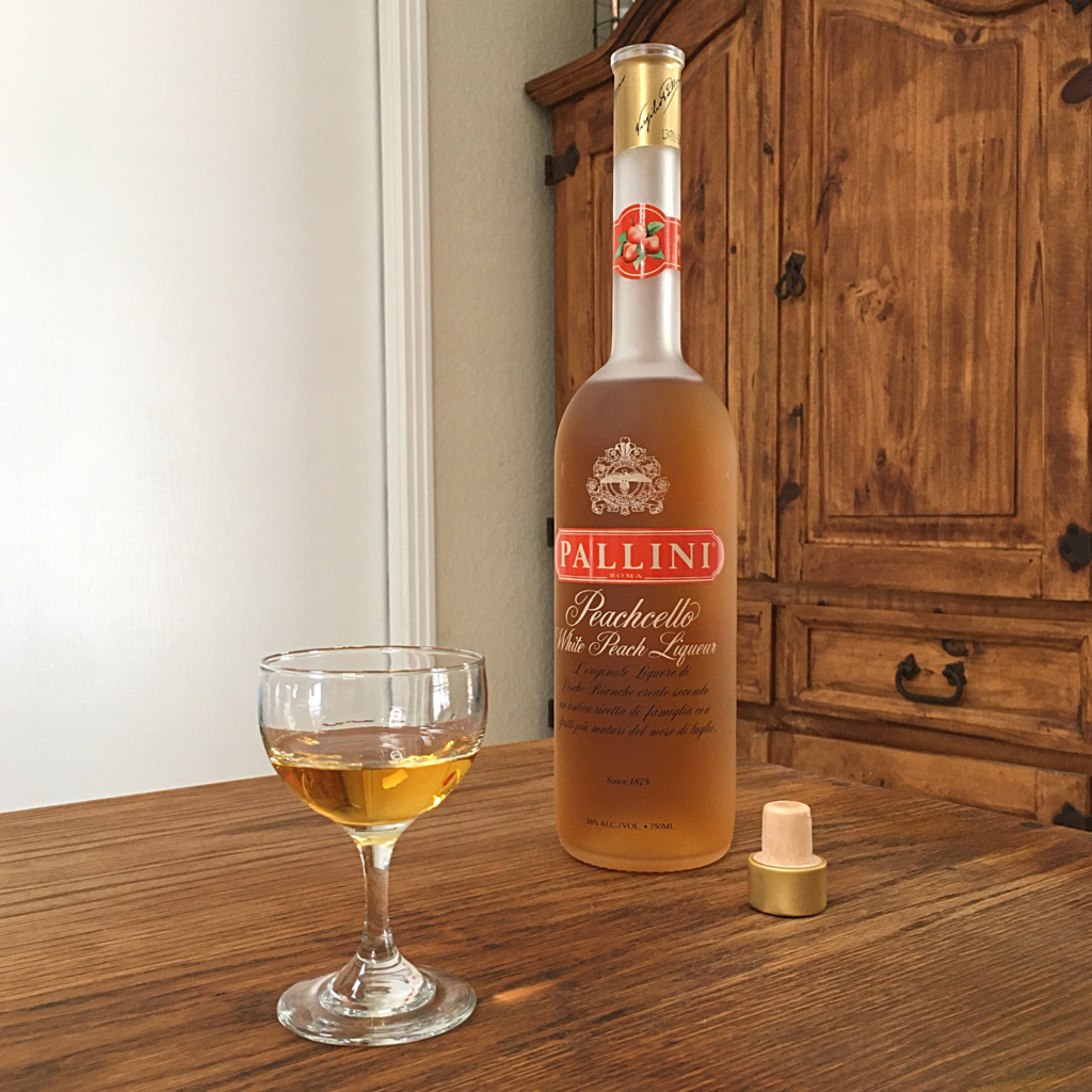 Tasting glass willed with orange liquid, next to an open bottle of Peachello White Peach Liqueur, sitting on a wooden table