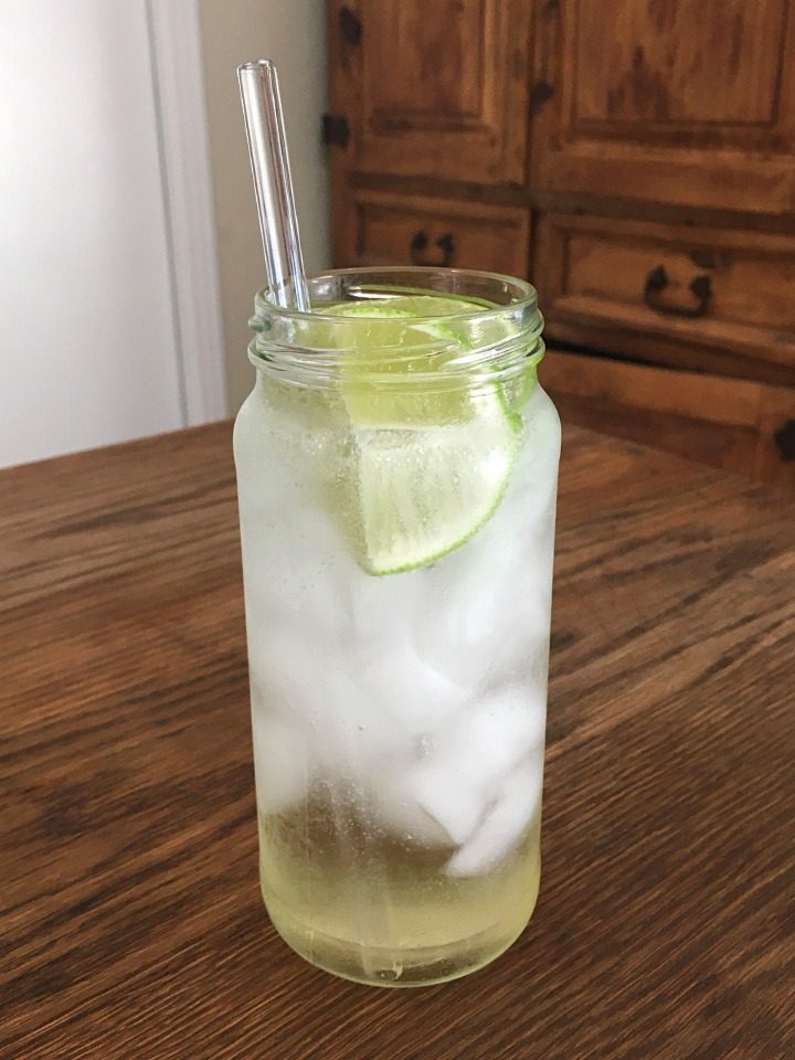 Light golden yellow cocktail in a tall glass with a clear glass straw and lime wedges, sitting on a wooden table