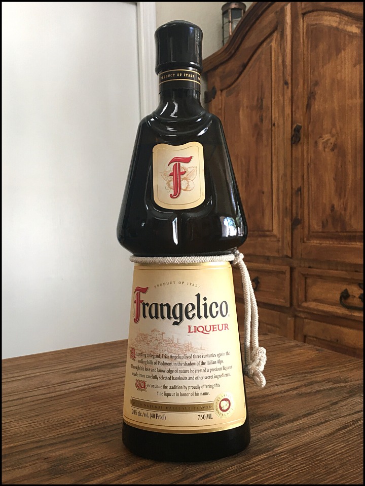 Tall bottle of Frangelico Hazelnut Liqueur sitting on a wooden table, with a mixed white and wooden background