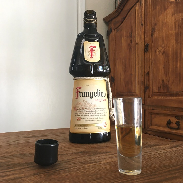 Open bottle of Frangelico Hazelnut Liqueur, next to a tall shot glass filled with light golden brown liquid, sitting on a wooden table