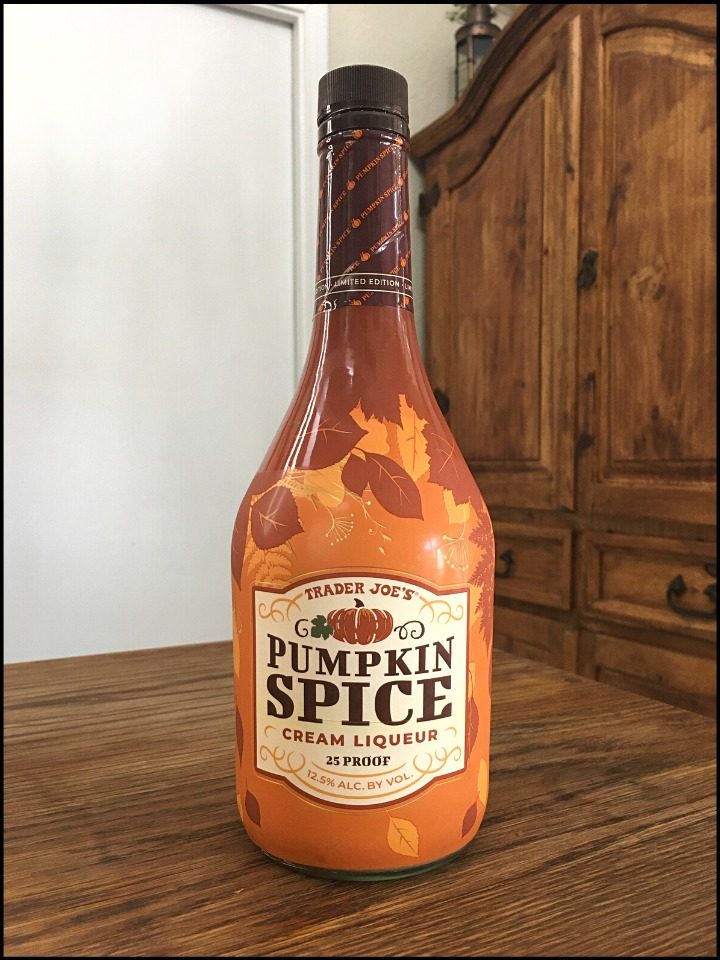 Bottle of Trader Joe's Pumpkin Spice Cream Liqueur sitting on a wooden table, in front of a mixed white and wood background