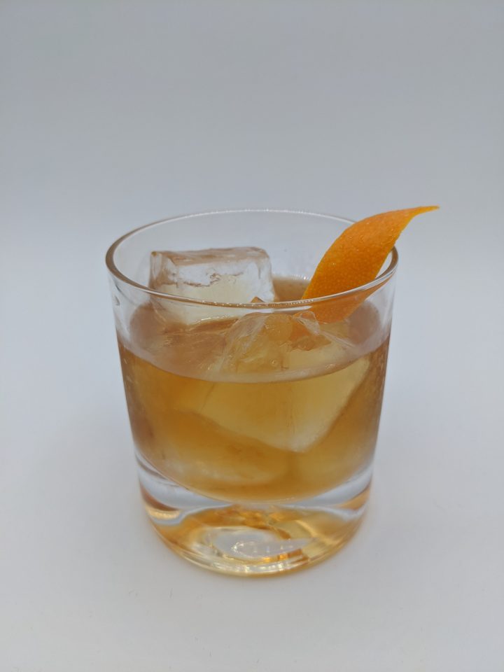 Gold liquid in a rocks glass with large cubes of ice with at orange peel garnish