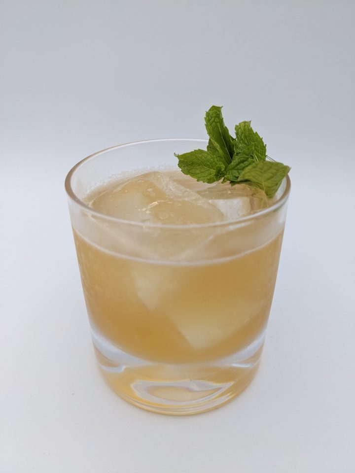 gold liquid with large ice cubes in a rocks glass with a sprig of mint as a garnish