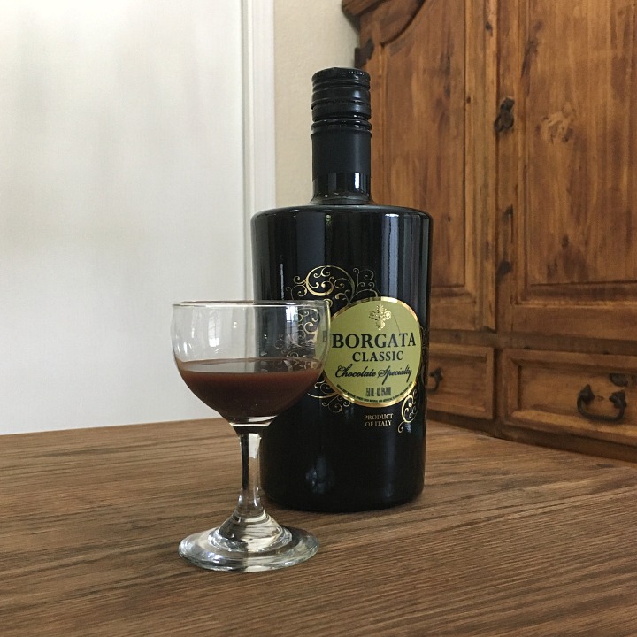Bottle of Borgata Chocolate Liqueur facing the righthand corner, slightly behind a small round glass filled with deep brown liquid, sitting on a wooden table