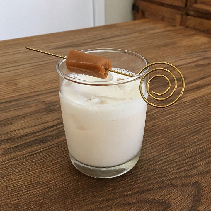Small cocktail glass filled with creamy white liquid on ice, with a gold cocktail pick and caramel garnish