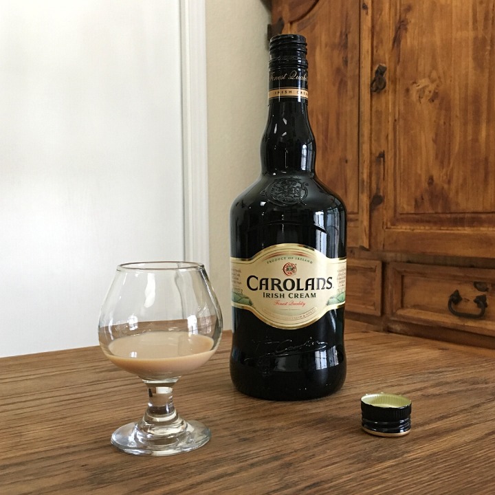 Open bottle of Carolans Irish Cream next to a small round glass with light beige liquid and a black bottle cap, all sitting on a wooden table