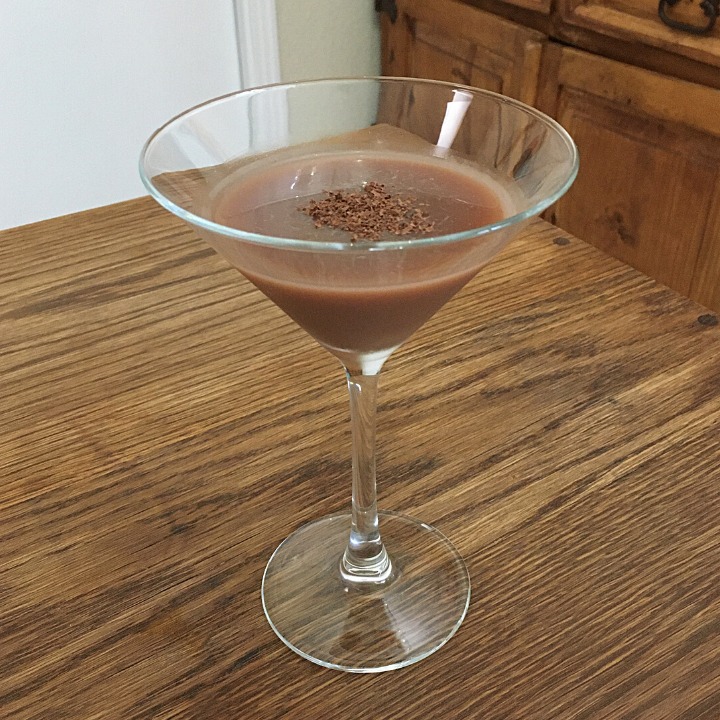 light brown cocktail with chocolate shavings garnish in a clear martini glass, sitting on a wooden table