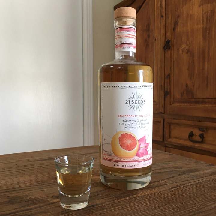 Bottle of 21 Seeds Grapefruit Hibiscus Tequila next to a clear shot glass filled with transparent pink liquid, both sitting on a wooden table