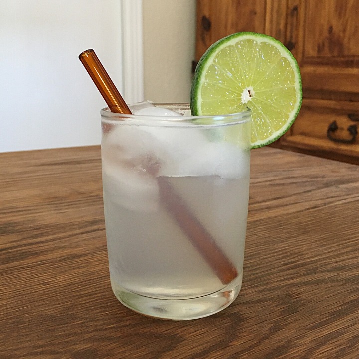 Translucent white cocktail with a lime wheel garnish and amber colored glass straw, sitting on a wooden table