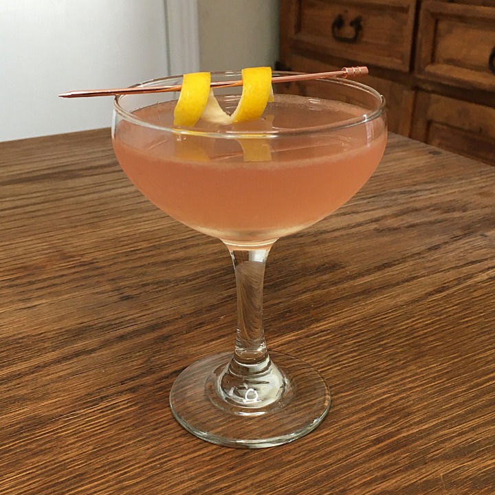 pink-orange cocktail in a coupe glass with a rose gold cocktail pick and lemon twist garnish, sitting on a wooden table