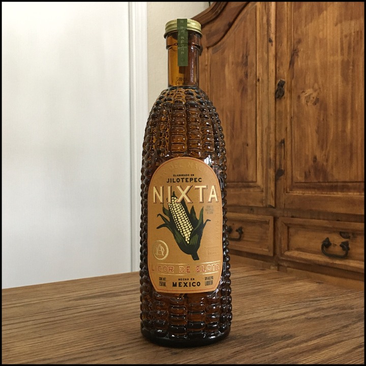 Bottle of Nixta Liqueur made with brown glass in a corn kernel textured design, sitting on a wooden table