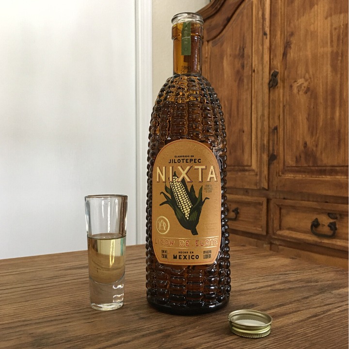 Open bottle of Nixta Liqueur next to a tall shot glass filled with light golden liquid and a golden bottle cap, all sitting on a wooden table