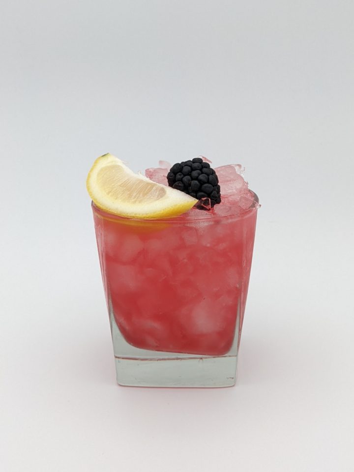 dark pink liquid in a cocltail glass with a blackberry and lemon wedge garnish