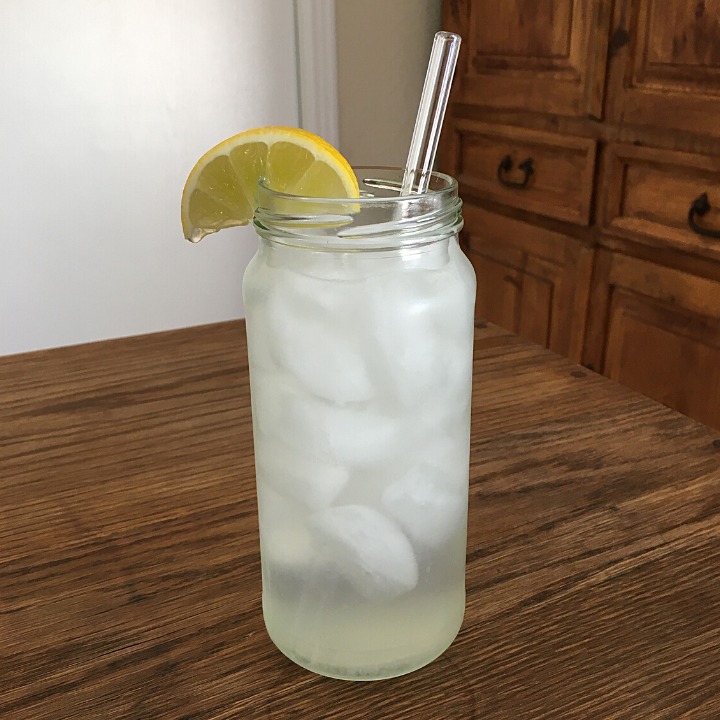 Tall jar glass filled with ice and translucent greenish-clear liquid, with a yellow lime garnish and a clear glass straw