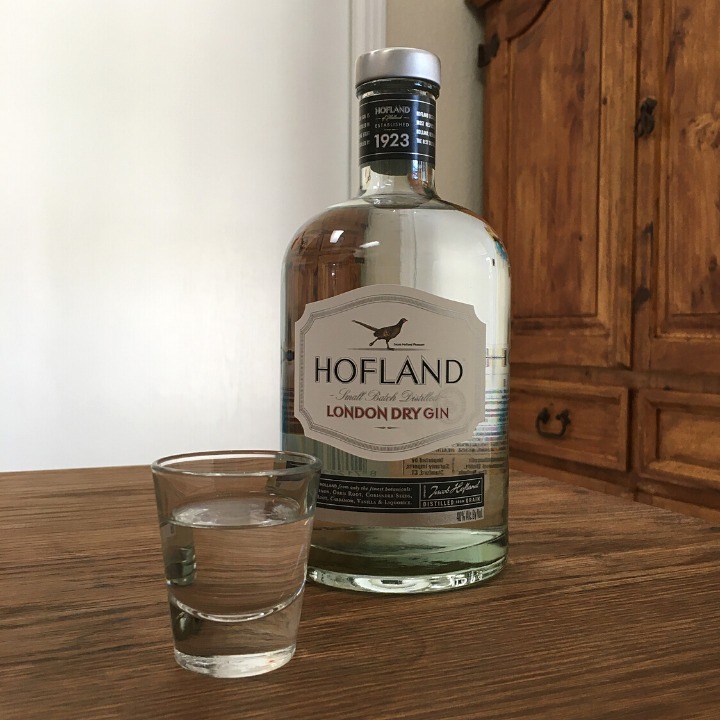 Bottle of Hofland Gin next to a shot glass filled with clear liquid, both sitting on a wooden table