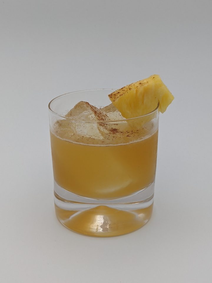 light brown liquid in a large rocks glass with large cubes of ice with a pineapple wedge garnish and a dusting of cinnamon